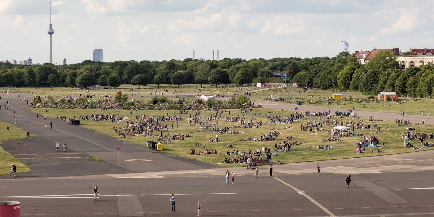 300 Hektar Grünfläche des Tempelhofer Felds. Viele Menschengruppen sitzen auf dem Rasen. Im Hintergrund ist die Skyline von Berlin sichtbar.